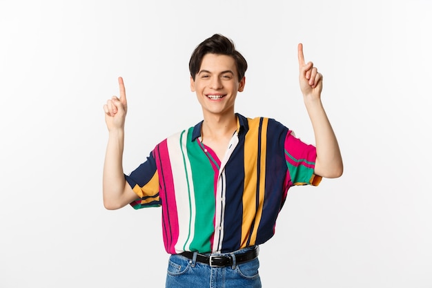 Homem bonito queer em roupas elegantes, mostrando o logotipo, apontando os dedos para cima e sorrindo satisfeito, em pé sobre o branco.