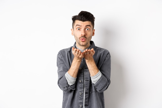 Homem bonito romântico soprando beijo no ar, parecendo bobo para a câmera, em roupas da moda em fundo branco.