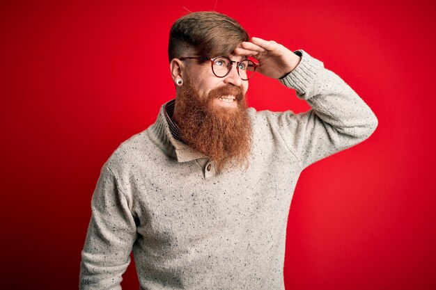 Homem bonito ruiva irlandesa com barba vestindo suéter casual e óculos sobre fundo vermelho muito feliz e sorrindo olhando para longe com a mão sobre a cabeça Conceito de pesquisa