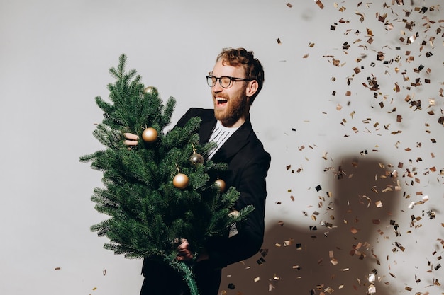 Homem bonito ruivo em uma jaqueta preta e óculos dança com uma árvore de Natal em suas mãos Conceito de festa de ano novo