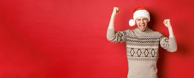 Homem bonito satisfeito, sentindo-se sortudo e feliz, gritando de alegria e fazendo punhos comemorando a vitória ou vitória, recebe um incrível presente de natal em pé no chapéu de papai noel sobre fundo vermelho