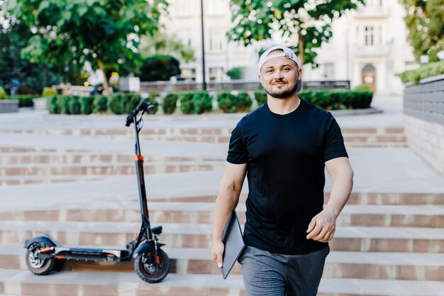 Homem bonito segurando laptop em sua mão em pé perto de scooter elétrico viajante de mochileiro de estilo de vida ativo