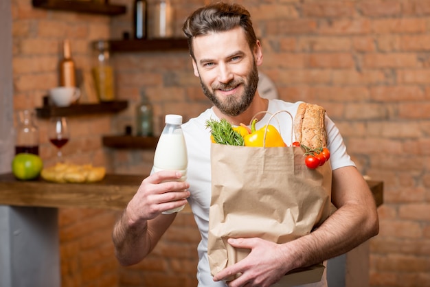 Homem bonito segurando leite com um saco de papel cheio de alimentos saudáveis na cozinha