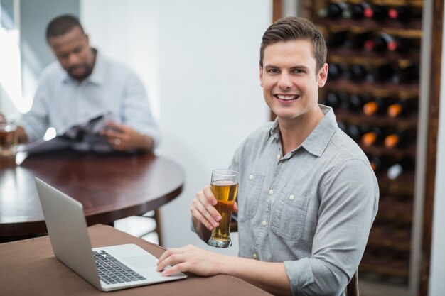 Homem bonito, segurando o copo de cerveja enquanto estiver usando o laptop