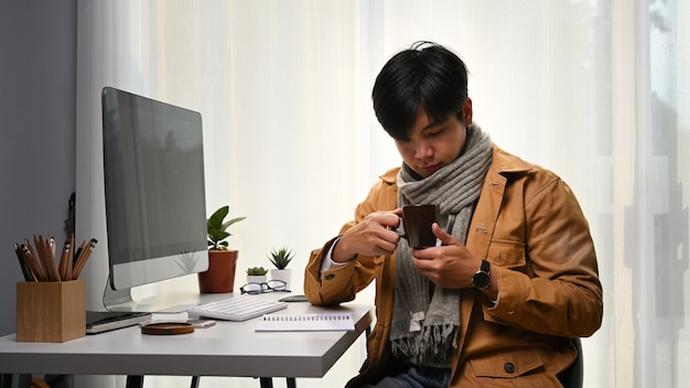 Homem bonito segurando uma xícara de café enquanto está sentado na frente do computador no escritório em casa.