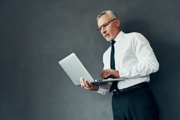 Homem bonito sênior com camisa elegante e gravata usando laptop em pé contra um fundo cinza