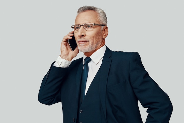 Homem bonito sênior em terno completo falando ao telefone em pé contra um fundo cinza