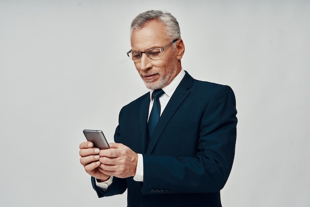 Homem bonito sênior em terno completo usando telefone inteligente e sorrindo em pé contra um fundo cinza