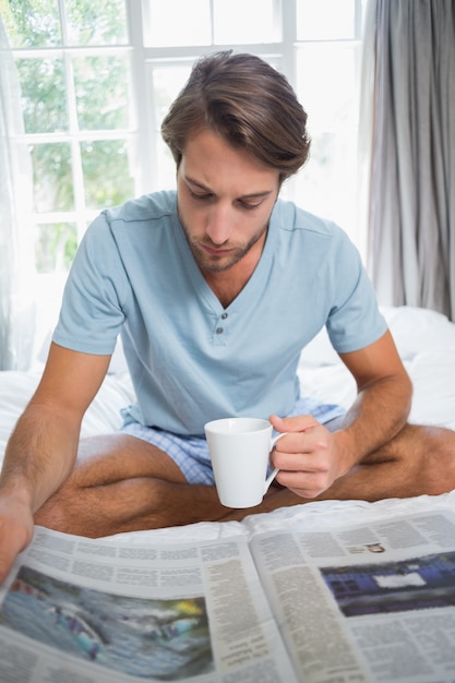 Homem bonito sentado na cama lendo o papel com café