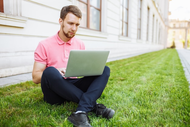 Homem bonito sentado na grama da cidade com um laptop, procura de emprego