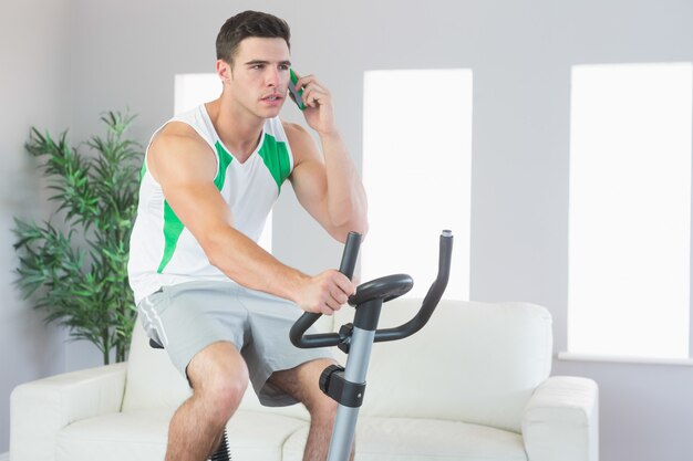 Homem bonito sério formação em bicicleta de exercício telefonando