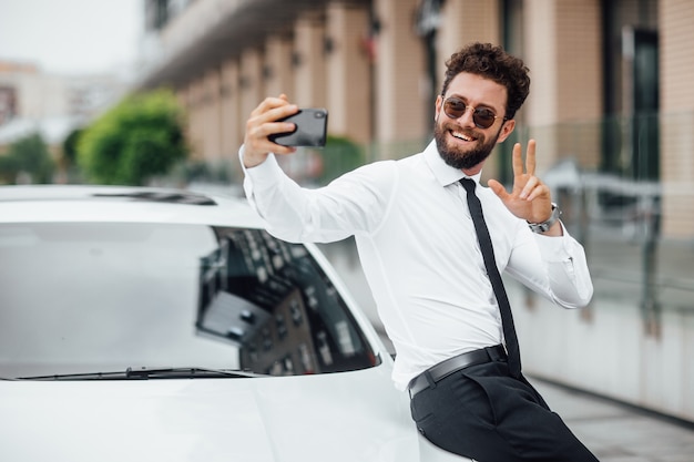 Homem bonito, sorridente e barbudo, de camisa branca, fazendo selfie perto de seu carro novo ao ar livre nas ruas da cidade