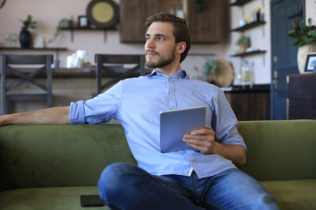 Homem bonito sorridente segurando e usando tablet com redes sociais em casa, freelancer feliz fazendo compras online, sentado no sofá aconchegante da sala de estar