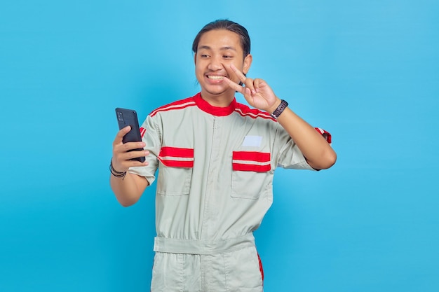 Homem bonito sorridente usando uniforme de mecânico fazendo videochamada e mostrando gesto de paz