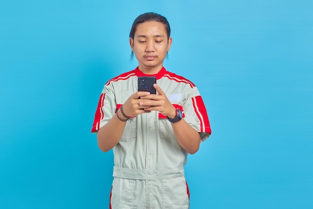 Homem bonito sorridente, vestindo uniforme mecânico, usando telefone celular isolado em fundo azul