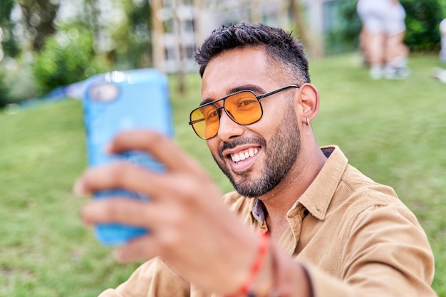 Homem bonito sorrindo para a câmera tendo uma selfie ao ar livre latino com óculos de sol da moda conversando em seu telefone celular