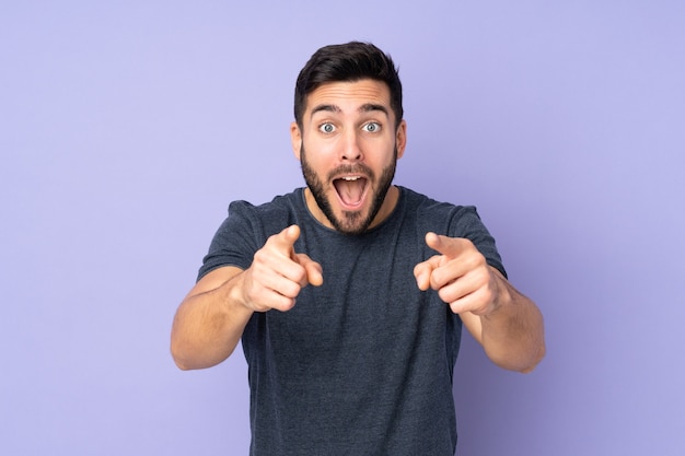 homem bonito surpreso e apontando para a frente sobre fundo roxo isolado