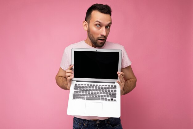 Homem bonito surpreso e espantado segurando um laptop olhando para a câmera em uma camiseta