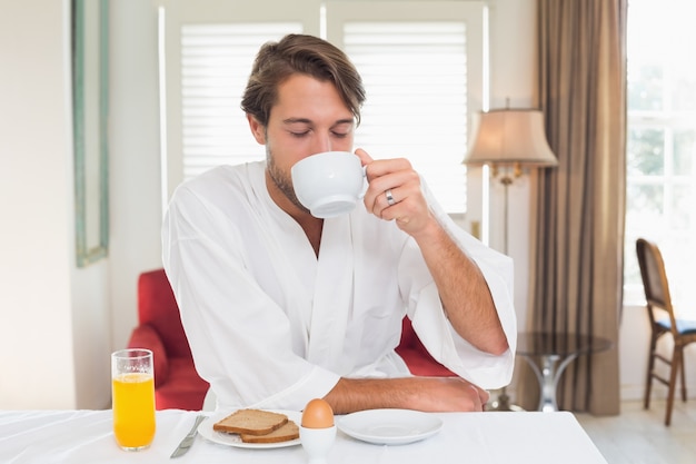 Homem bonito tomando café da manhã em seu roupão bebendo café