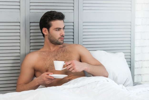 Homem bonito tomando café na cama olhando para longe