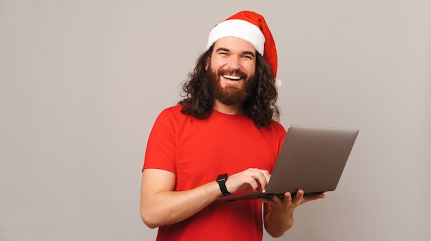 Homem bonito usando chapéu de Natal está segurando um laptop enquanto sorri largo