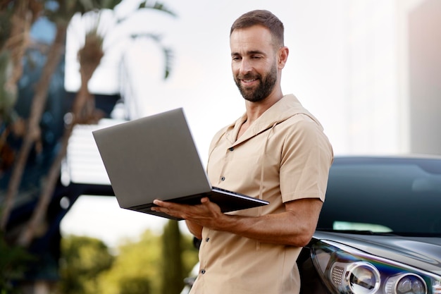 Homem bonito usando computador portátil fazendo diagnóstico de carro. sorrindo freelancer latino trabalhando online