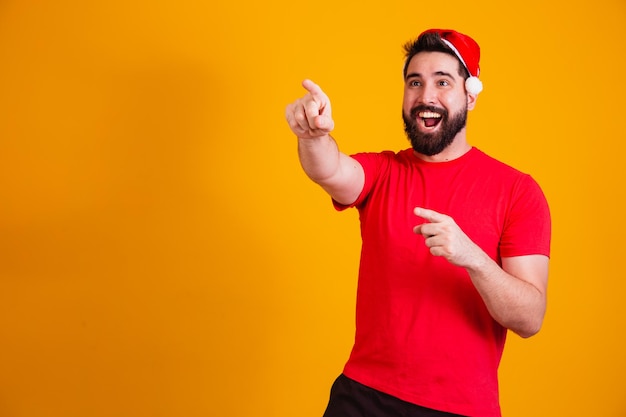 Homem bonito vestido para o Natal com chapéu de Papai Noel, apontando para o espaço de texto livre. promoção de fim de ano