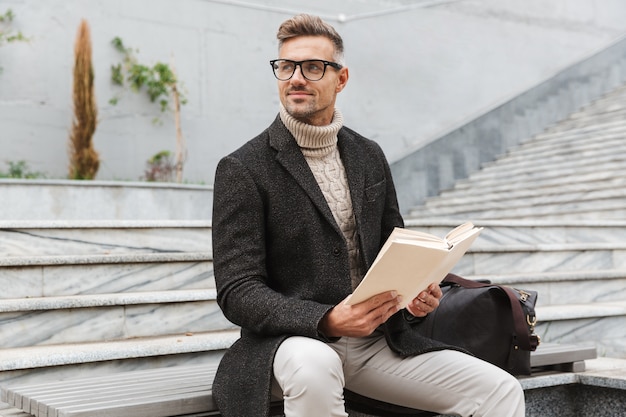 Homem bonito vestindo jaqueta lendo um livro sentado ao ar livre