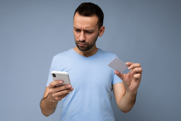 Homem bonito vestindo roupas do dia a dia isoladas na parede do fundo, segurando e usando telefone e crédito