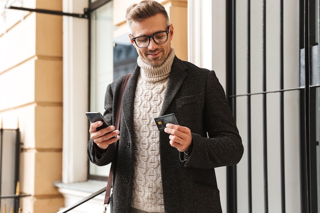 Homem bonito vestindo um casaco, caminhando ao ar livre, fazendo compras online com o celular e o cartão de crédito
