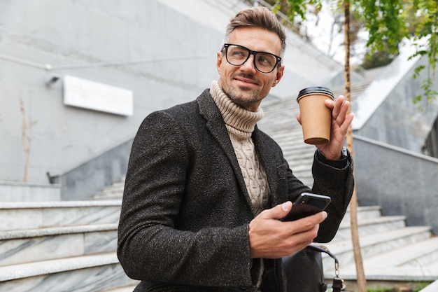 Homem bonito vestindo uma jaqueta segurando um telefone celular, bebendo café para viagem enquanto está sentado ao ar livre
