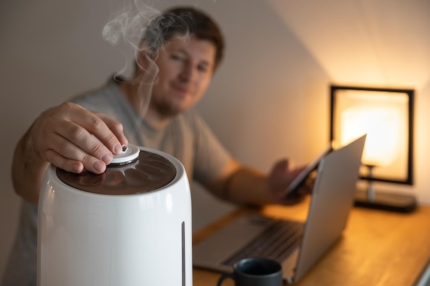 Homem bonito vira o bico do umidificador de ar branco. umidificação do ar seco. foco seletivo no vapor. conceito de pureza do ar. aumentando a umidade da sala. macho trabalhando no laptop. fechar-se.