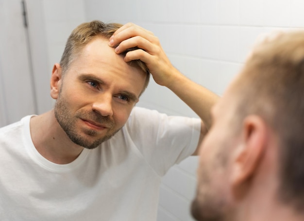 Homem branco caucasiano de meia idade com barba curta olha para o cabelo no espelho no banheiro e preocupado com a calvície O conceito do problema da perda de cabelo masculino calvície precoce e alopecia