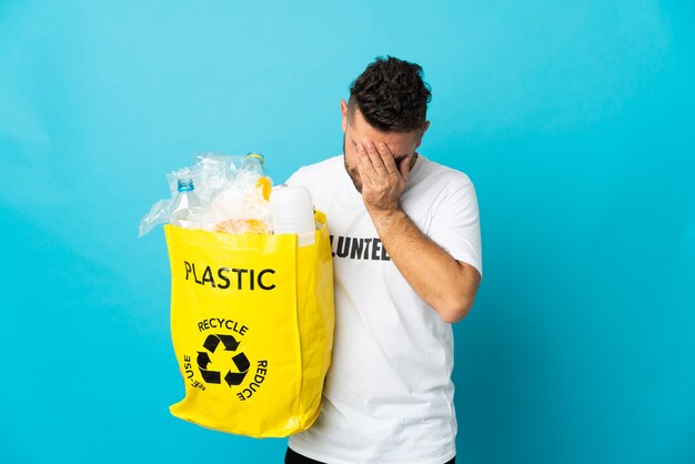 Homem branco segurando uma sacola cheia de garrafas plásticas para reciclar isoladas em um fundo azul com expressão cansada e doente