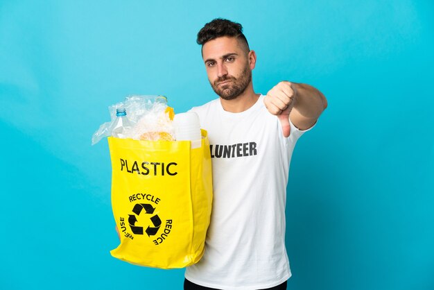 Homem branco segurando uma sacola cheia de garrafas plásticas para reciclar isoladas em um fundo azul, mostrando o polegar para baixo com expressão negativa