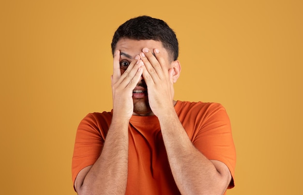 Homem brasileiro assustado em camiseta laranja cobrindo o rosto com as mãos em pé sobre fundo amarelo do estúdio