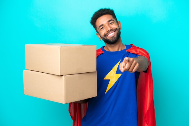 Homem brasileiro de entrega de super-herói isolado em fundo azul, apontando para a frente com expressão feliz