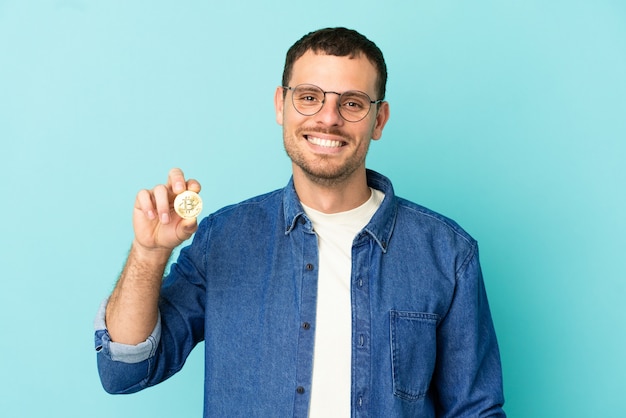 Homem brasileiro segurando um Bitcoin sobre um fundo azul isolado, posando com os braços na cintura e sorrindo