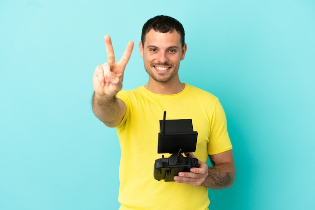 Homem brasileiro segurando um controle remoto de drone sobre fundo azul isolado, sorrindo e mostrando sinal de vitória