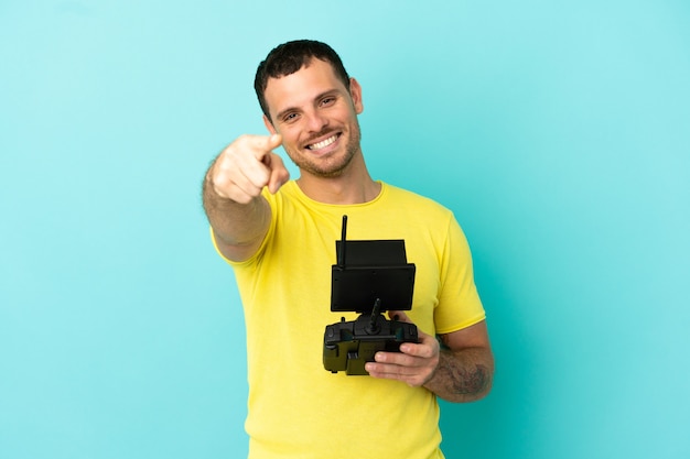 Homem brasileiro segurando um controle remoto de drone sobre um fundo azul isolado apontando para a frente com uma expressão feliz