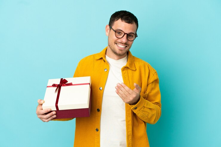 Foto homem brasileiro segurando um presente sobre fundo azul isolado, convidando para vir com a mão. feliz que você veio