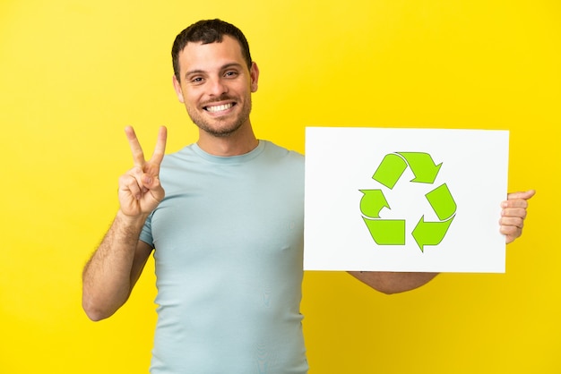 Homem brasileiro sobre fundo roxo isolado segurando um cartaz com o ícone de reciclagem e comemorando uma vitória