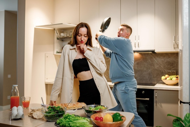 Homem brincando balançando frigideira em direção à cabeça de uma mulher desavisada enquanto eles cozinham juntos na cozinha moderna em casa