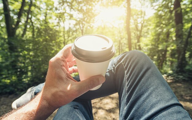 Homem brindando café para levar a xícara contra a luz solar explosiva na floresta. Tiro do ponto de vista