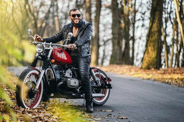 Homem brutal barbudo de óculos escuros e jaqueta de couro, sentado em uma motocicleta na estrada na floresta