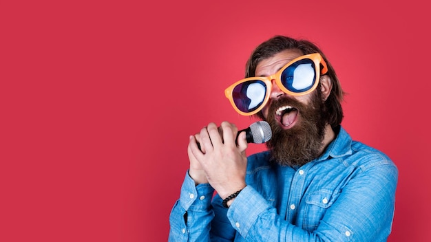 Homem brutal barbudo engraçado com bigode usa óculos de festa e canta música no microfone karaokê