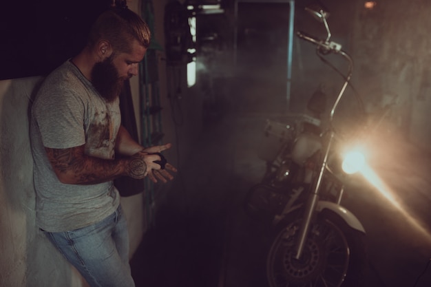 Homem brutal bonito com uma barba está de pé em sua garagem no contexto de uma motocicleta e olhando para longe