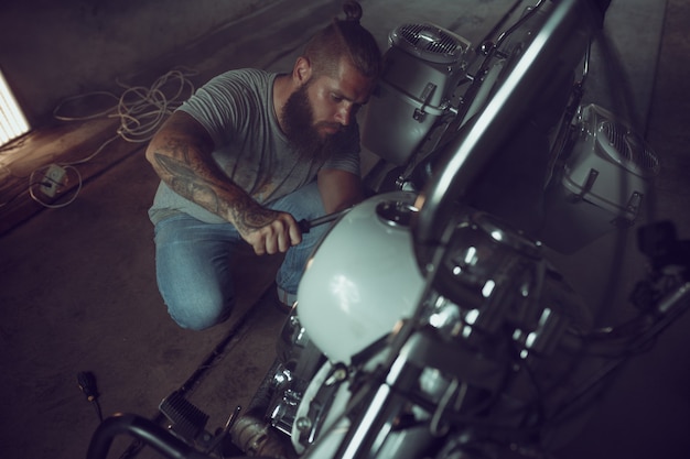 Homem brutal bonito com uma barba que repara uma motocicleta em sua garagem