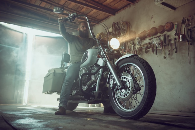 Homem brutal bonito com uma barba, sentado em uma motocicleta em sua garagem