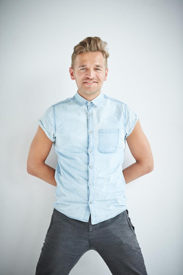 Foto homem brutal com uma barba pequena está encostado na parede vestindo uma camisa azul de manga curta e calça cinza as mãos estão atrás das costas foto de alta qualidade